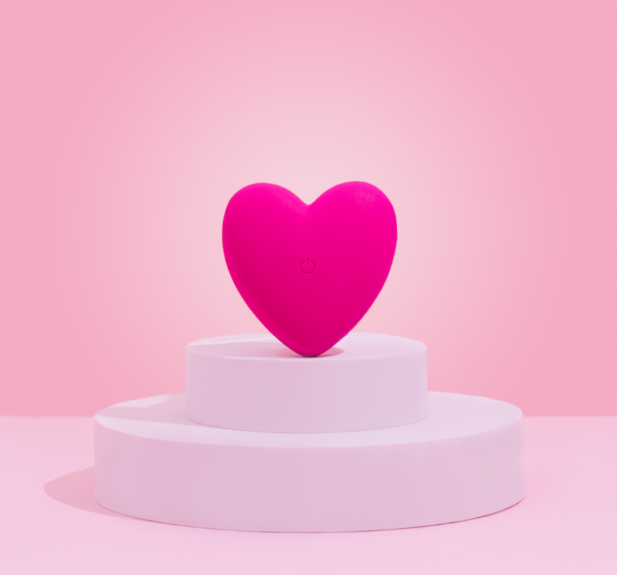 A pink heart-shaped vibrator placed on a white stand against a pink background